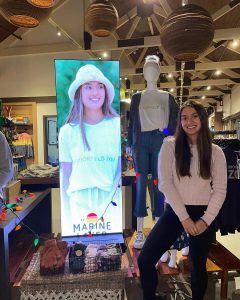 Abby in a gift shop seated next to a digital screen display of her modeling in the campaign for Marine Layer