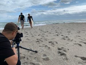 Roy behind the camera filming two surfers on the beach about to enter the water